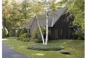Dark wood-sided single-story building surrounded by a manicured lawn, trimmed shrubs, and trees including a birch in the foreground.
