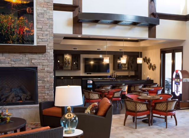 A cozy common area with a stone fireplace on the left, a seating arrangement with cushioned chairs and a glass table lamp in the foreground, and a dining area with round tables and red cushioned chairs in the background. Pendant lights hang above the dining area, and a jukebox is visible near glass doors on the right.