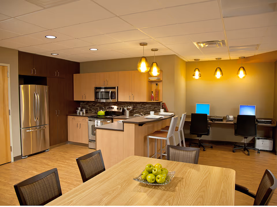 A modern kitchen and workspace area with wooden cabinets, stainless steel refrigerator, stove, and microwave. There is a kitchen island with two white bar stools and place settings. In the foreground, a wooden table with a bowl of green apples and four chairs is visible. The workspace area has two computer stations with black chairs and pendant lights hanging from the ceiling.
