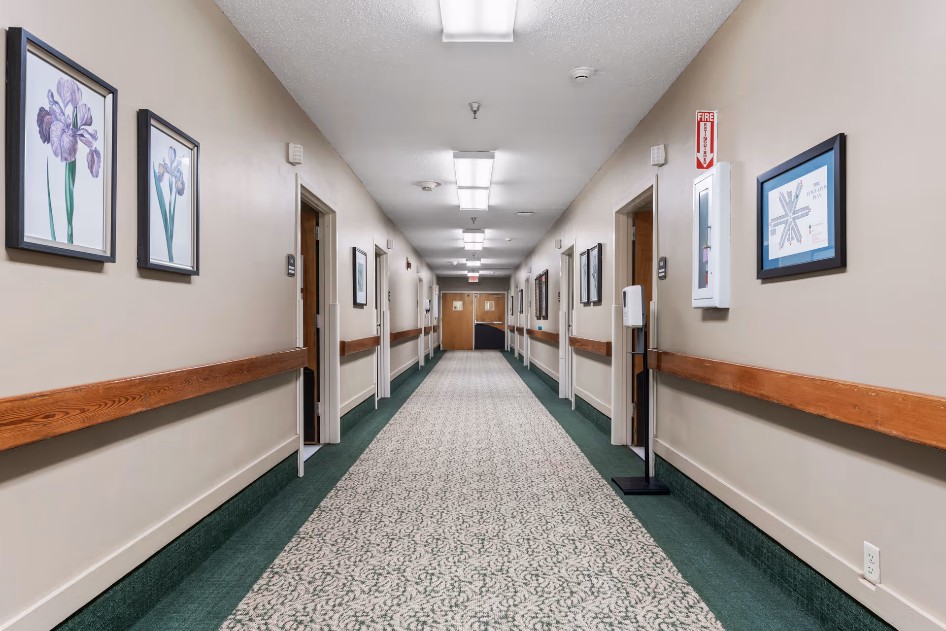A long, well-lit hallway in a nursing and care center with beige walls, green and patterned carpet, wooden handrails on both sides, framed floral artwork on the walls, several open doors leading to rooms, and a fire extinguisher cabinet on the right wall.