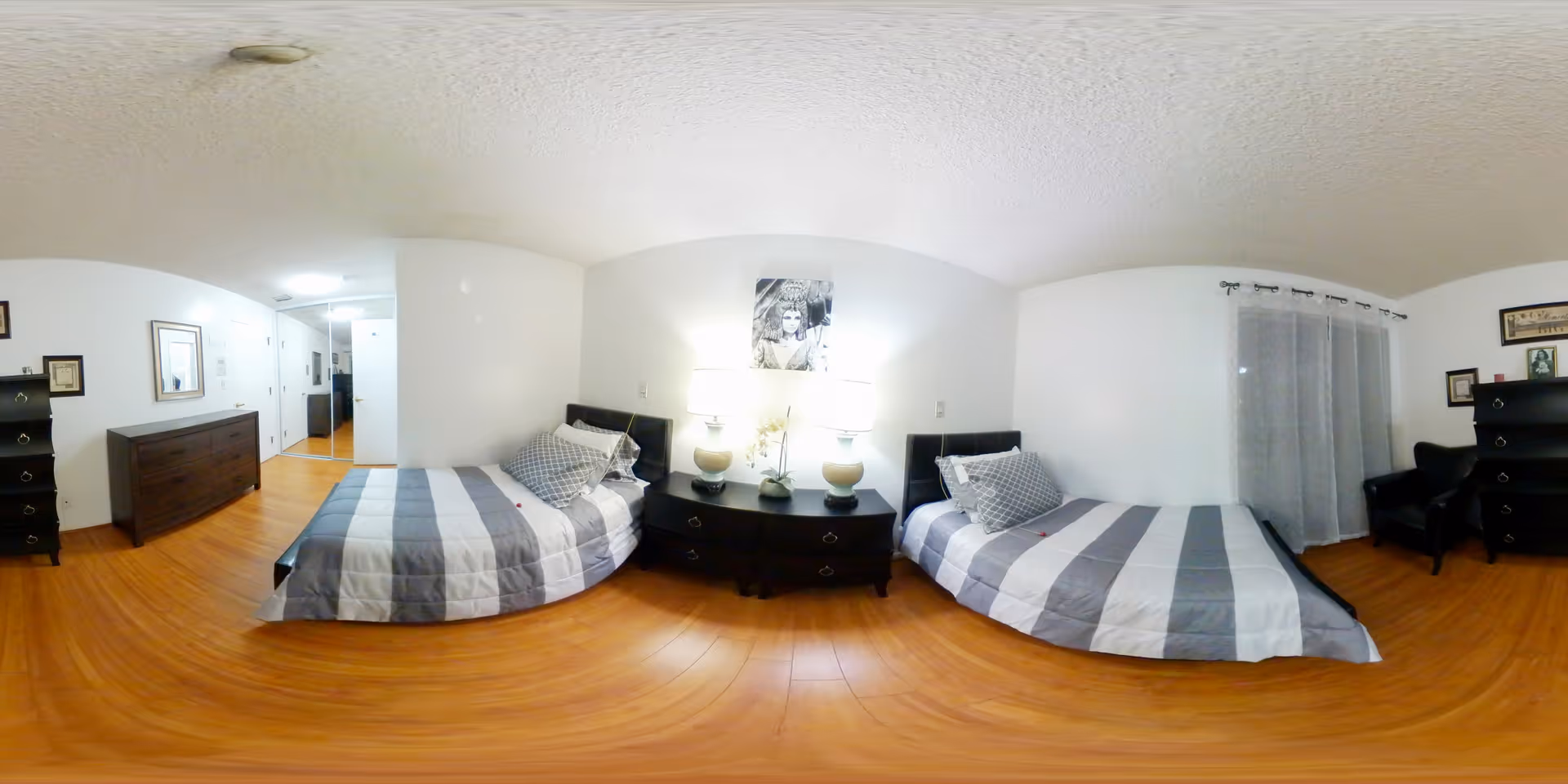Bright bedroom with two twin beds separated by a nightstand with lamps, dressers, and hardwood floors.