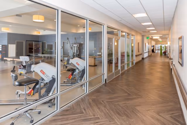 A long hallway with wood-patterned flooring and white walls, featuring large glass windows on the left side that reveal a fitness room with exercise equipment inside. The hallway is well-lit with ceiling lights and has framed pictures on the walls.