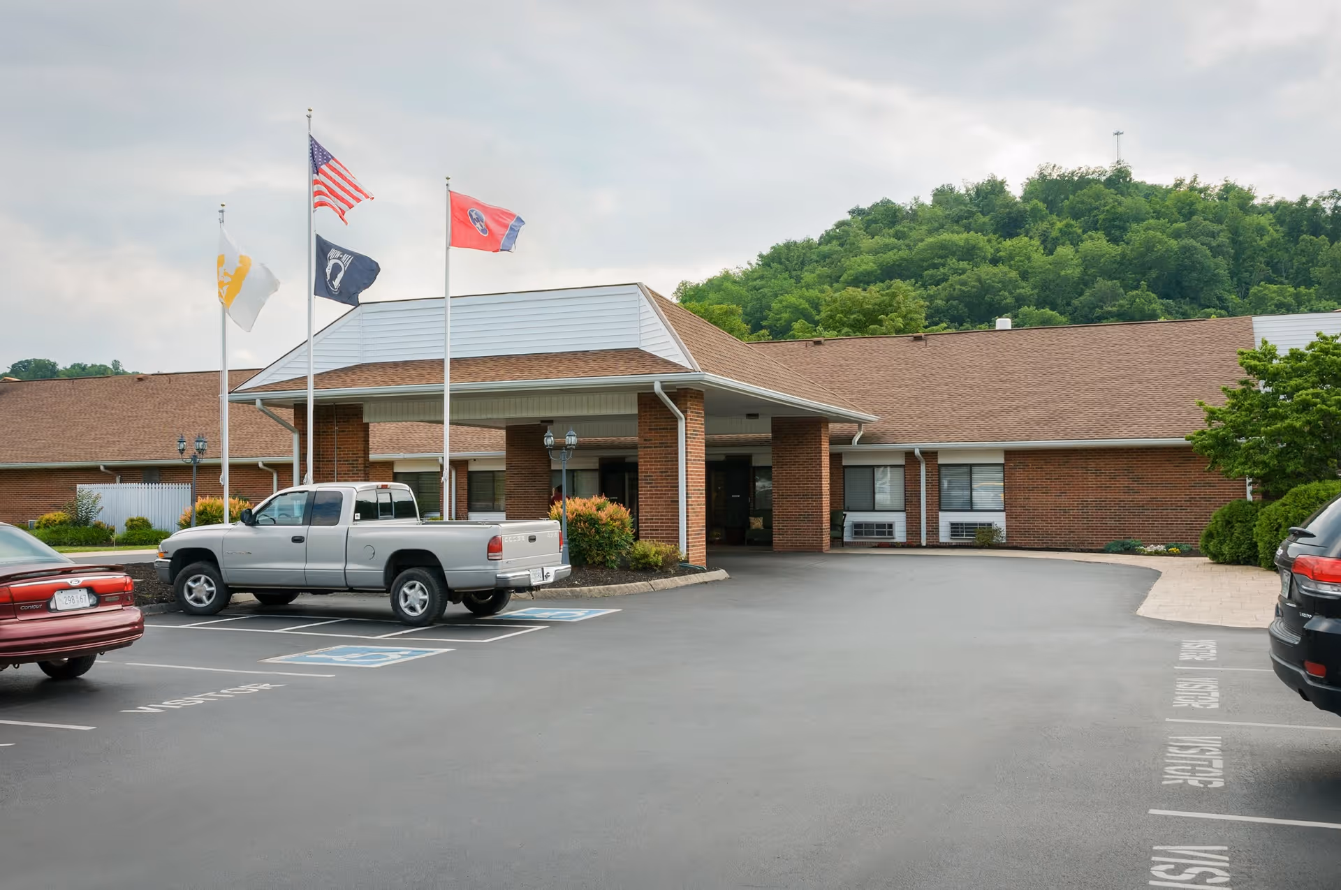 Front exterior view of a single-story brick building with a covered entrance, three flagpoles displaying flags, a parking lot with several parked vehicles, and a green wooded hill in the background under a cloudy sky.