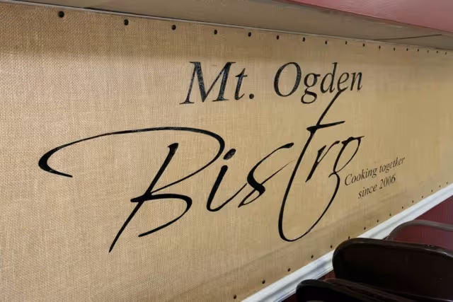 A burlap-textured wall sign reading 'Mt. Ogden Bistro' with 'Cooking together since 2006' displayed above dining chairs.