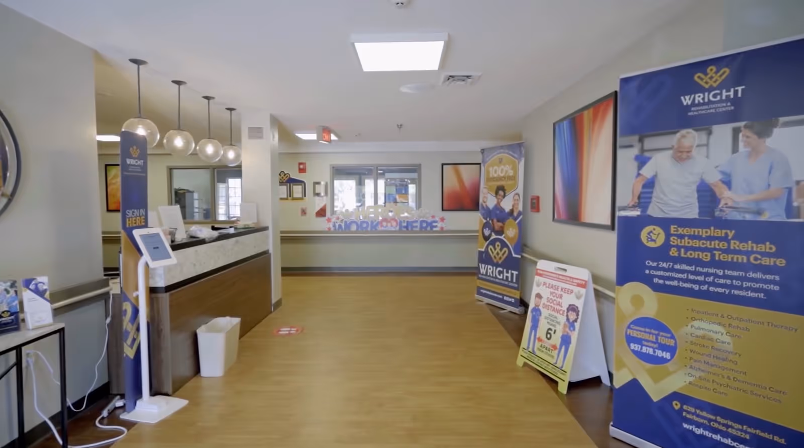 Interior view of a healthcare facility reception area with a wooden floor, a reception desk on the left, informational banners and signs about rehabilitation and long-term care on the right, and a glass door at the far end. The space is well-lit with ceiling lights and pendant lights above the desk.