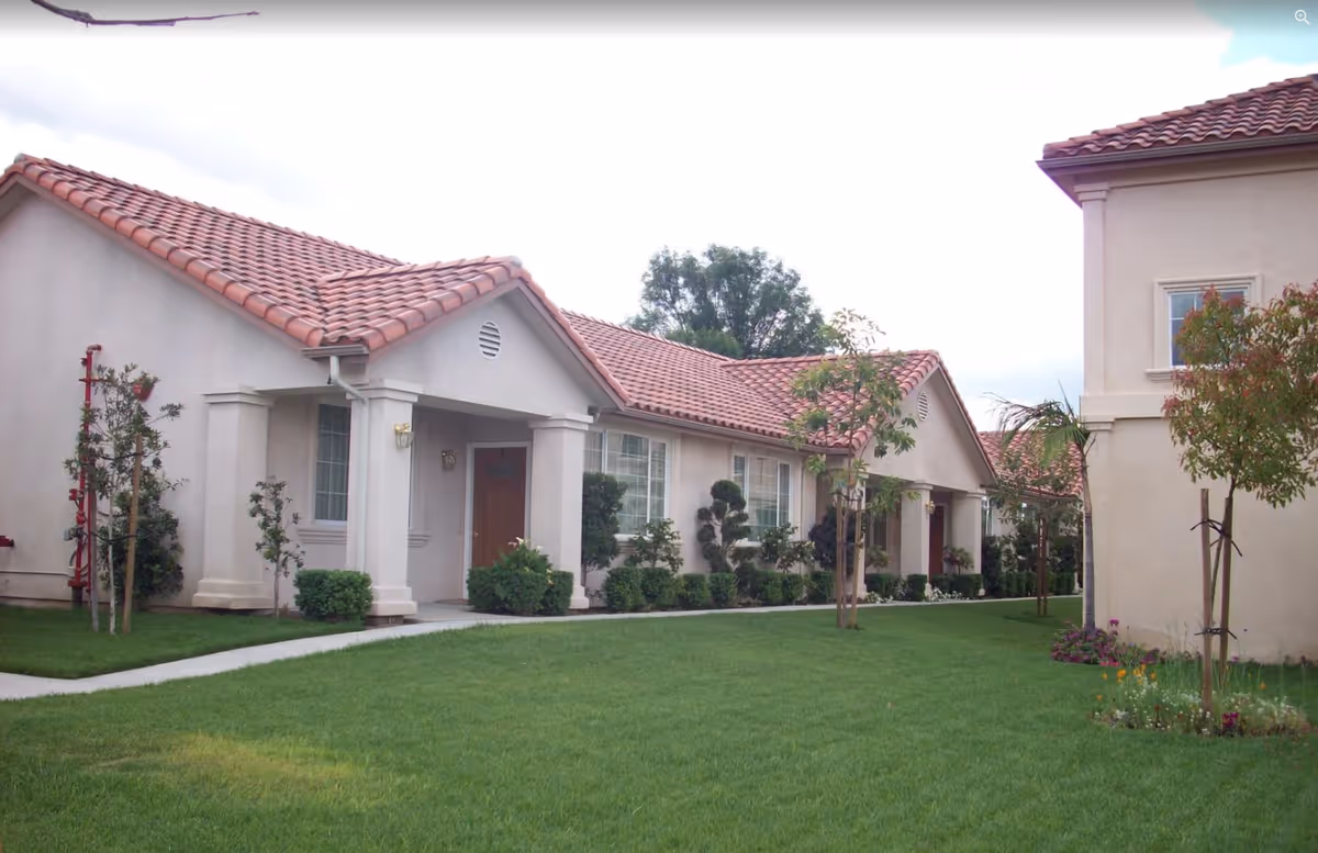 Exterior view of a retirement center building with beige walls and red tiled roofs, surrounded by a well-maintained green lawn and small trees and shrubs.