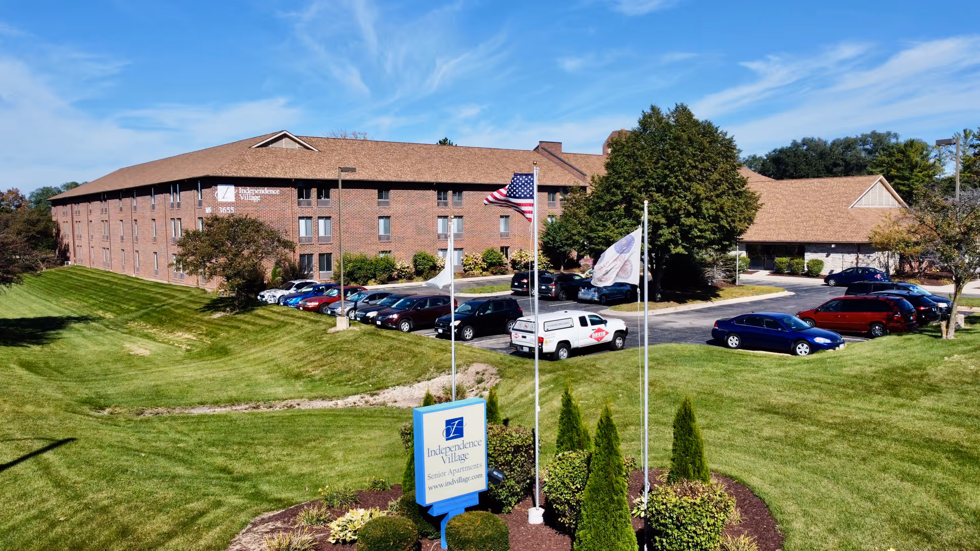Exterior view of Independence Village senior living facility showing a large brick building with multiple windows, a parking lot with several cars, three flagpoles with flags, and a landscaped grassy area with shrubs and trees under a blue sky.