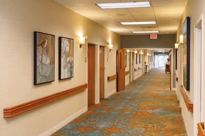 A well-lit hallway in a senior living facility with patterned carpet, beige walls, wooden handrails, and several closed doors. The hallway is decorated with framed paintings and has wall-mounted lights. An exit sign is visible at the far end.