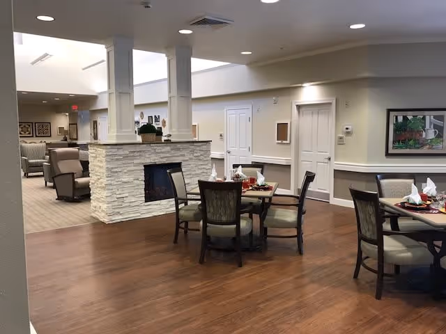 Interior view of a senior living facility dining area with wooden floors, round tables set with napkins and tableware, cushioned chairs, and a stone fireplace in the center. The background shows a lounge area with armchairs and soft lighting.