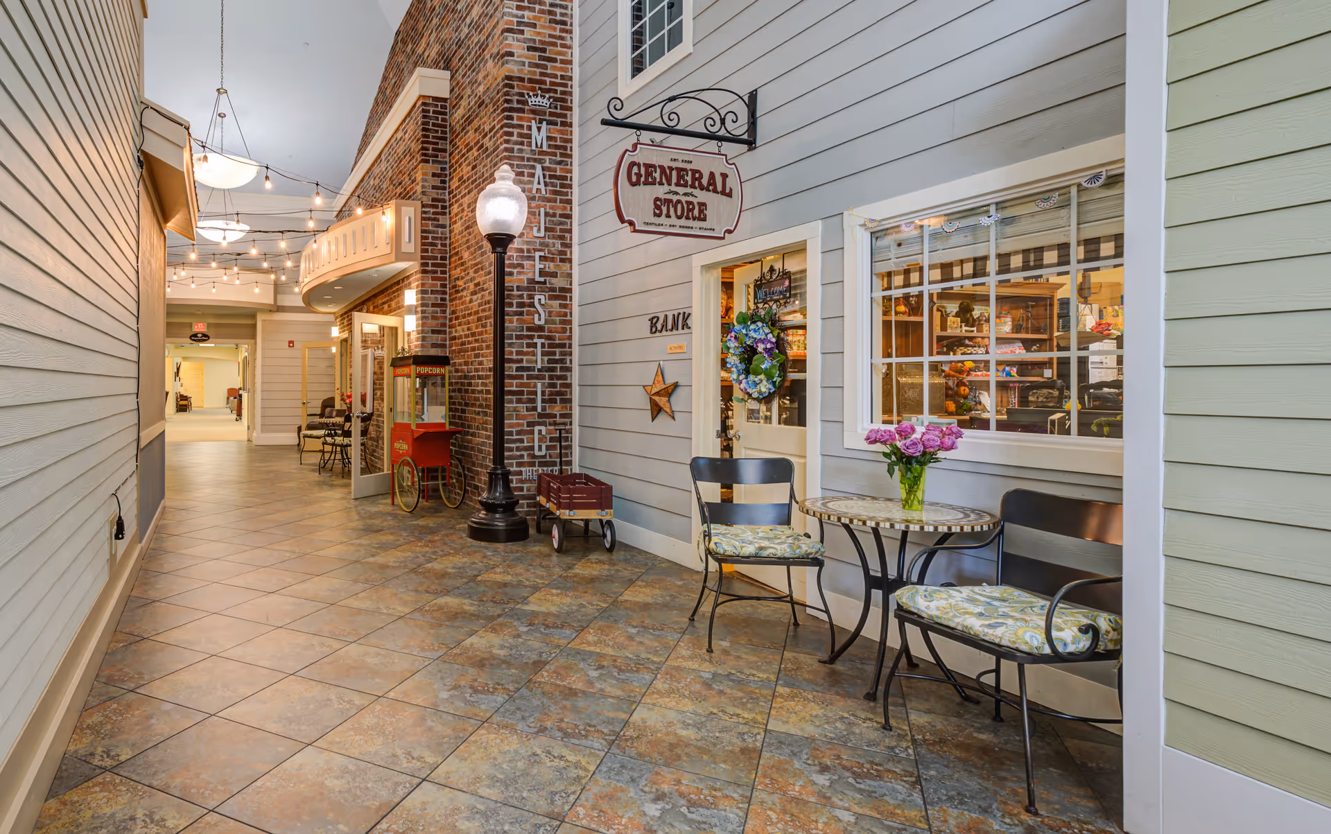 Indoor hallway designed to look like a small town street with a General Store and Bank storefronts. There is a small table with two chairs and a vase of purple flowers on the right side. String lights hang from the ceiling, and a popcorn machine and a street lamp are visible along the hallway.