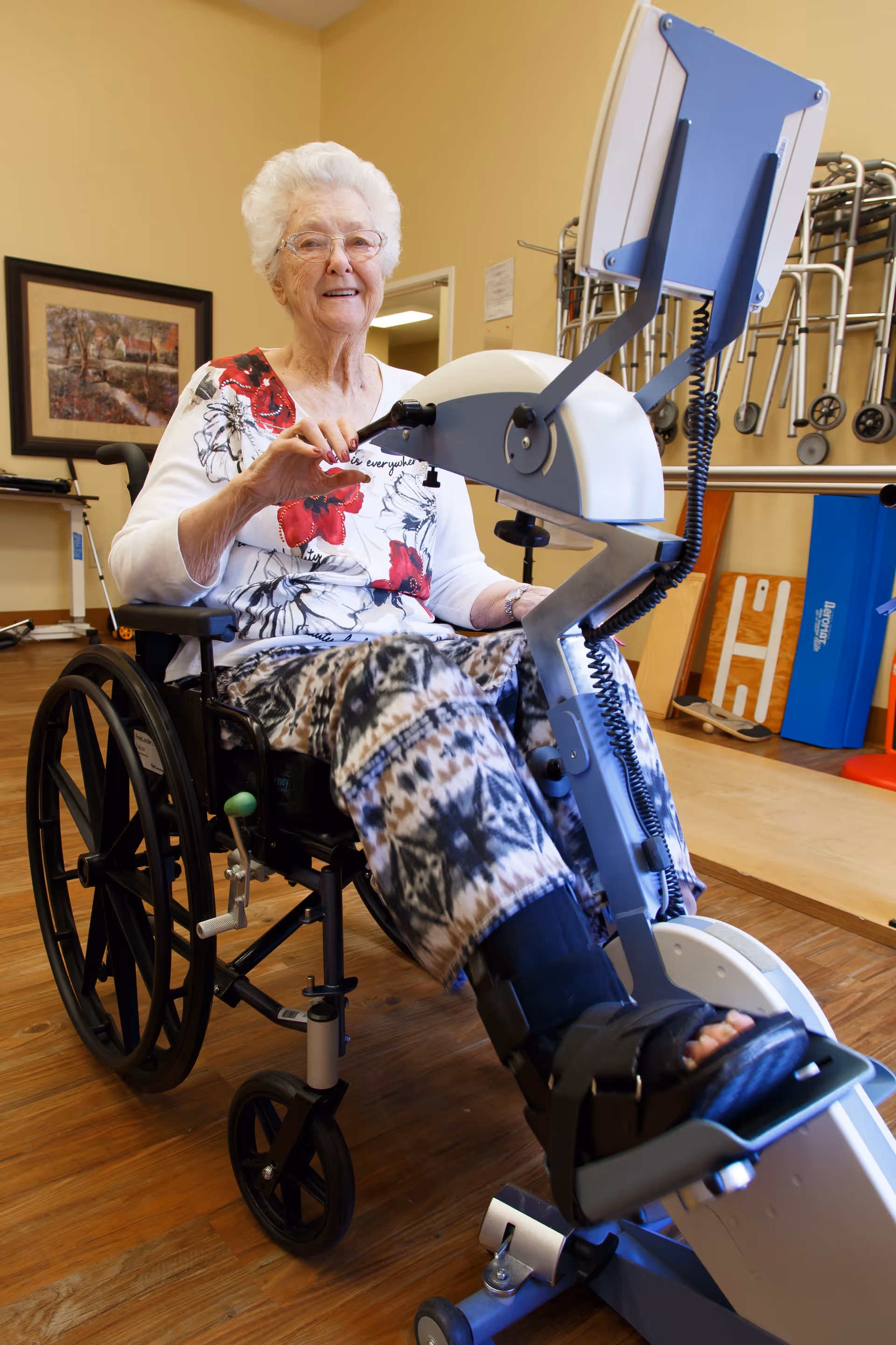 An elderly woman sitting in a wheelchair using a leg exercise machine in a rehabilitation or therapy room. The room has wooden floors, a framed picture on the wall, and various mobility aids like walkers in the background.