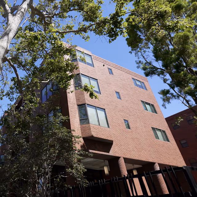 Exterior view of a multi-story brick building with several windows, surrounded by green trees under a clear blue sky.