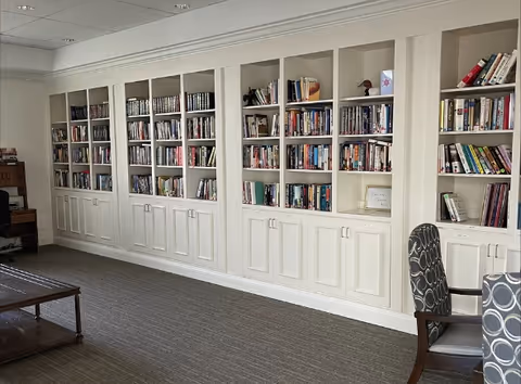 Built-in white bookshelves filled with books line a carpeted common room with chairs and a coffee table.