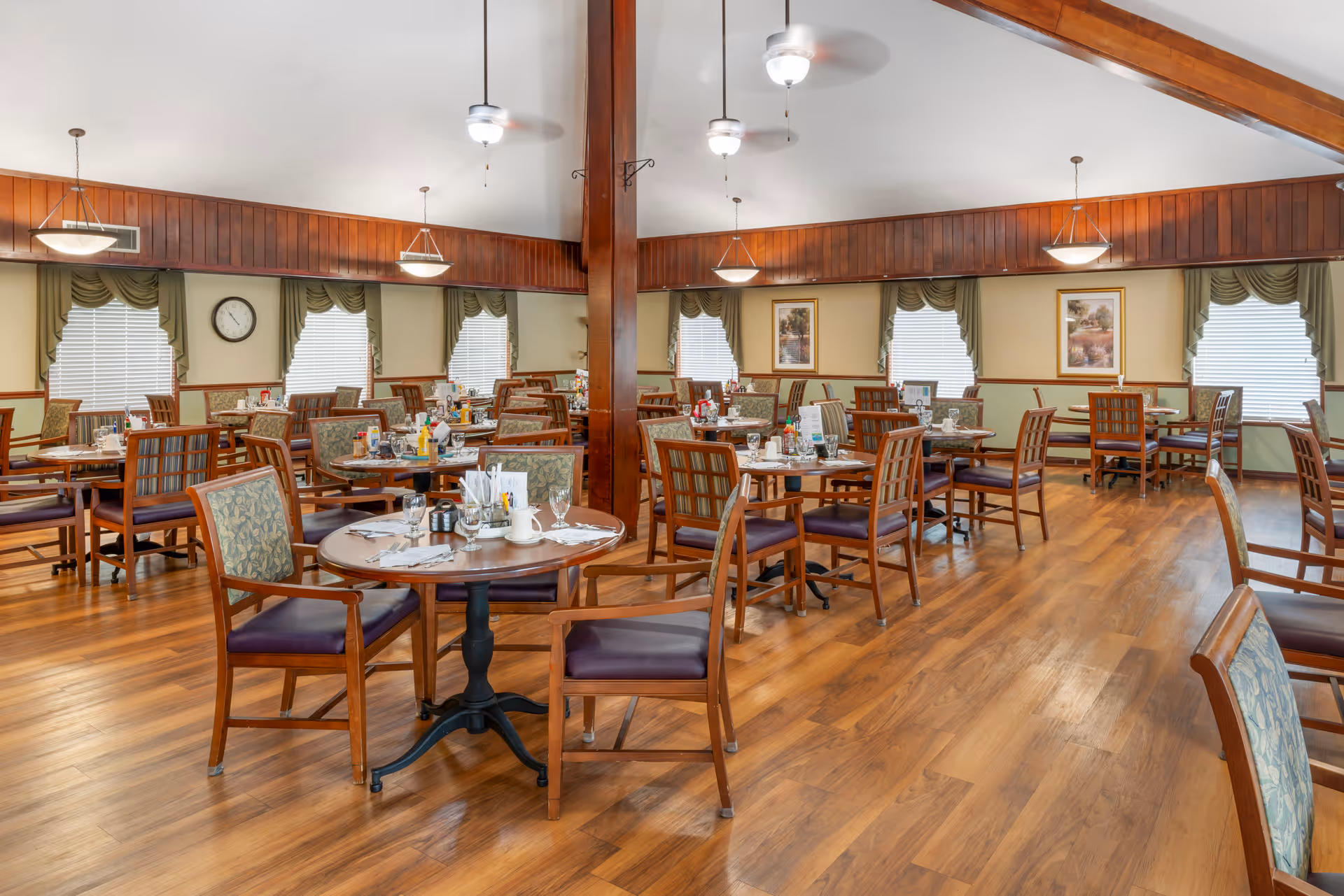 Spacious dining room with multiple round and rectangular wooden tables surrounded by chairs with patterned upholstery. The room has wooden floors, green walls with wooden paneling, several windows with green valances, ceiling fans, and hanging light fixtures. Tables are set with glasses, napkins, condiments, and menus.