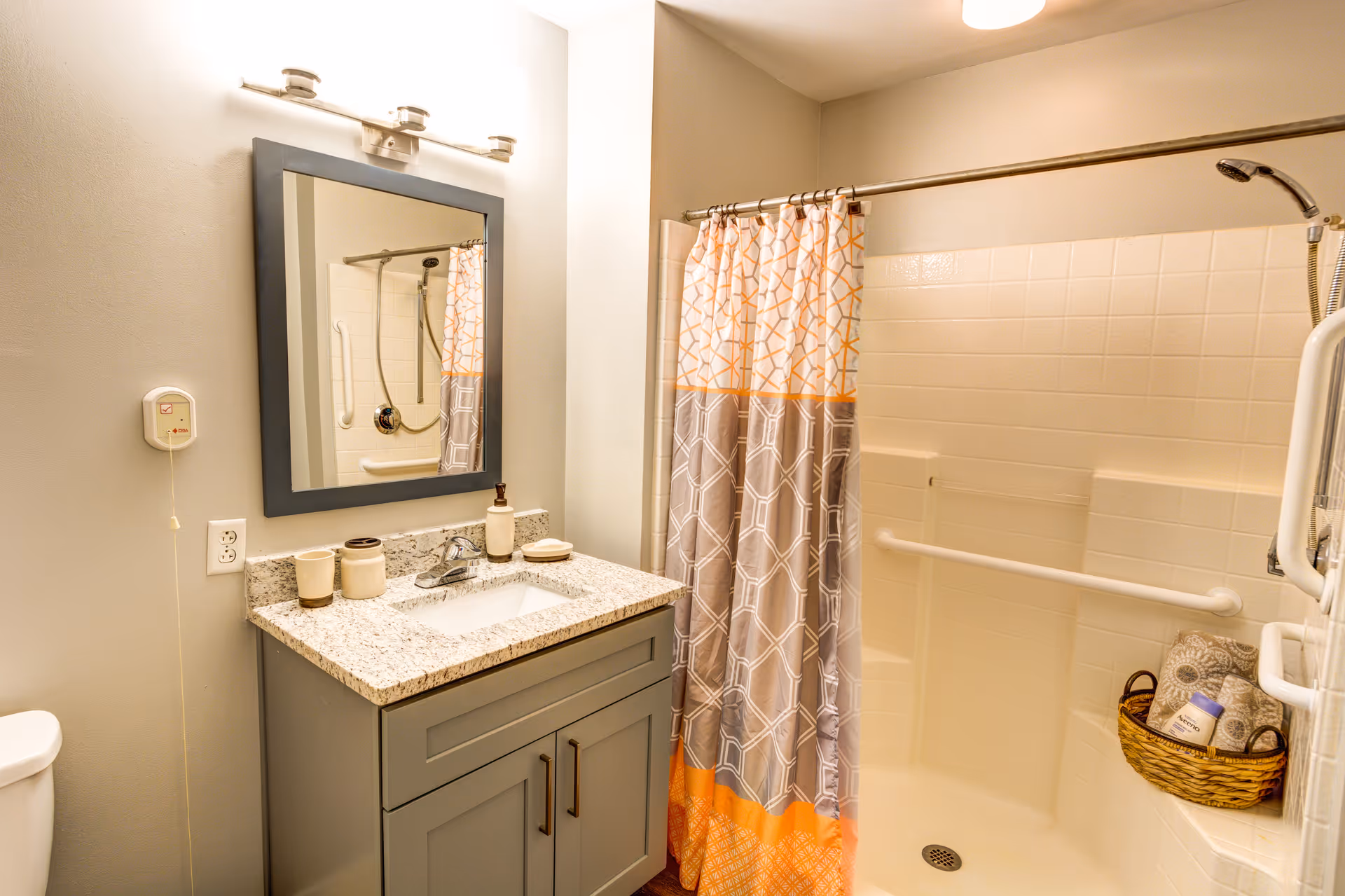 A bathroom with a gray vanity topped with a granite countertop and a rectangular mirror above it. The shower area has white tiled walls, a grab bar, and a shower curtain with orange and gray geometric patterns. A basket with towels and toiletries is placed on a built-in shower seat.