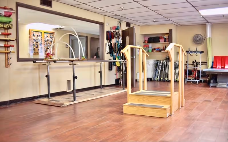 Indoor rehabilitation room with parallel bars, wooden practice steps, and assorted mobility aids and equipment on shelves and racks.