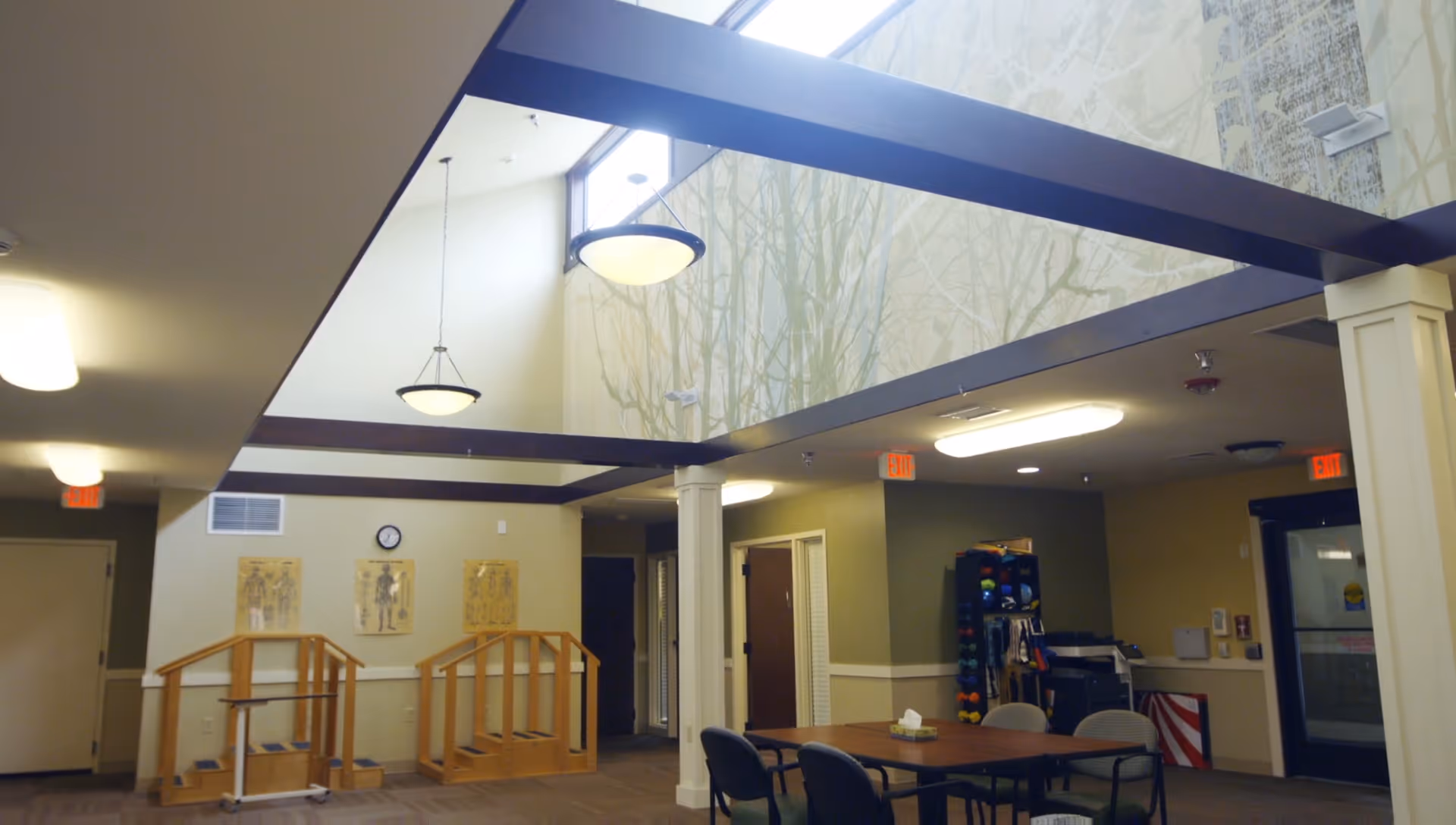 Interior view of a senior living facility common area with high ceilings and large windows. The room features wooden handrails for physical therapy or exercise, a table with chairs, exercise equipment, and wall decorations including anatomical charts and a clock.