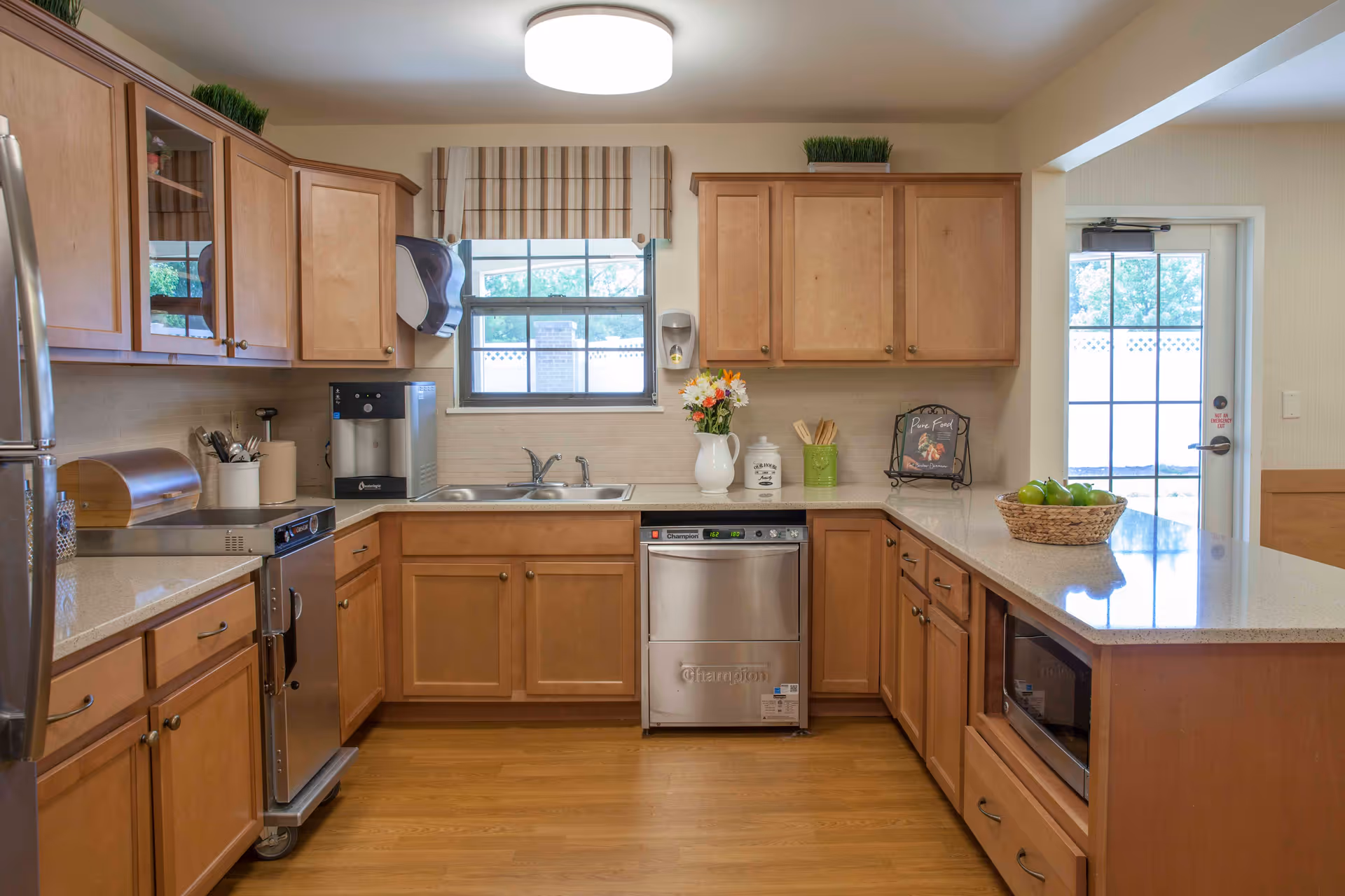 A clean and well-lit kitchen with wooden cabinets, a stainless steel refrigerator, dishwasher, and microwave. There is a double sink under a window with a striped valance, a countertop with a basket of green apples, and various kitchen utensils and decorations including a vase with flowers and a small framed sign.