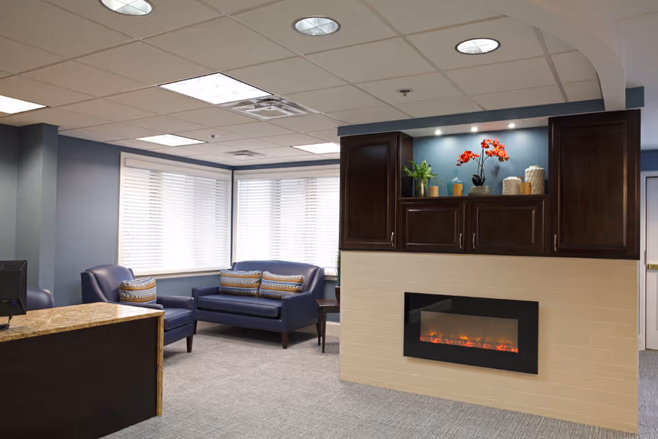 A cozy interior lounge area with a modern electric fireplace set in a beige tiled wall. Above the fireplace are dark wooden cabinets decorated with plants and vases. To the left, there is a reception desk with a granite countertop and a computer monitor. In the background, there are two blue armchairs and a blue loveseat with patterned cushions, positioned near large windows with white blinds. The walls are painted blue and the ceiling has recessed lighting.