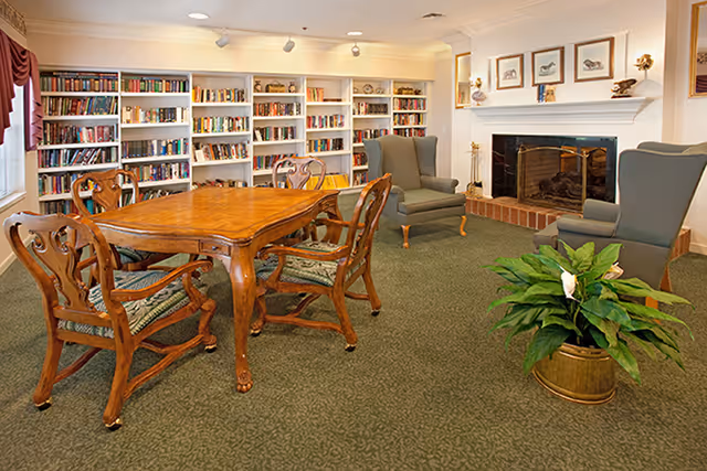 A cozy common area with a wooden table and four carved wooden chairs with patterned cushions. Behind the table is a large bookshelf filled with books. To the right, there are two green upholstered armchairs facing a brick fireplace with a white mantel decorated with framed pictures and small sculptures. A green potted plant in a brass container is placed on the carpeted floor in the foreground.