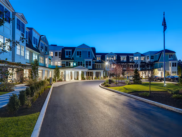 Exterior view of The Mariner Marblehead senior living facility at dusk, showing a large multi-story building with many windows, a curved driveway, landscaped greenery, and an American flag on a flagpole.
