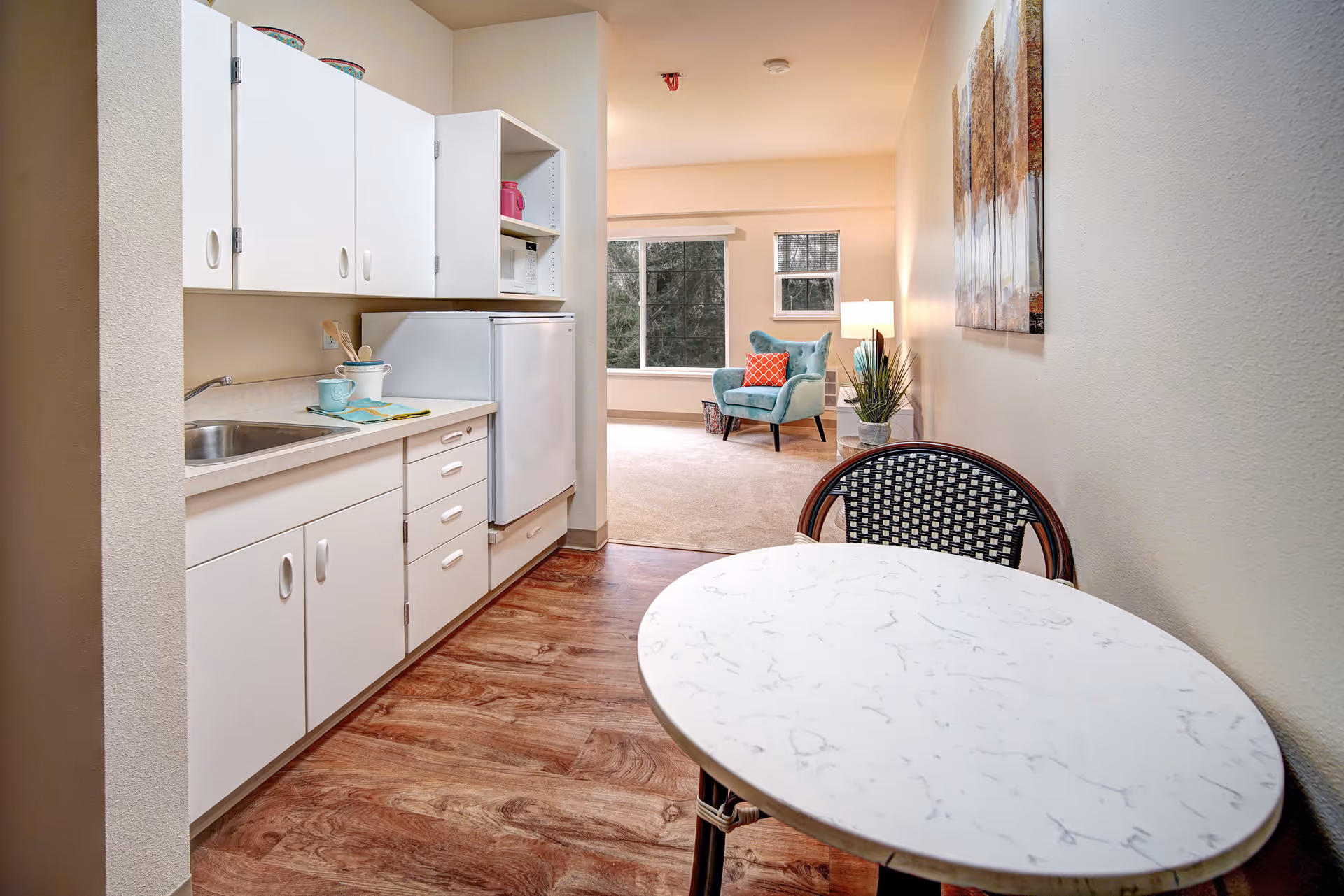 A small kitchen area with white cabinets, a countertop with a sink, and a white refrigerator. In the foreground, there is a round table with a black and white woven chair. Beyond the kitchen, a living area is visible with a teal armchair, a red patterned pillow, a floor lamp, and a large window letting in natural light. The floor has wood-like flooring in the kitchen and carpet in the living area.