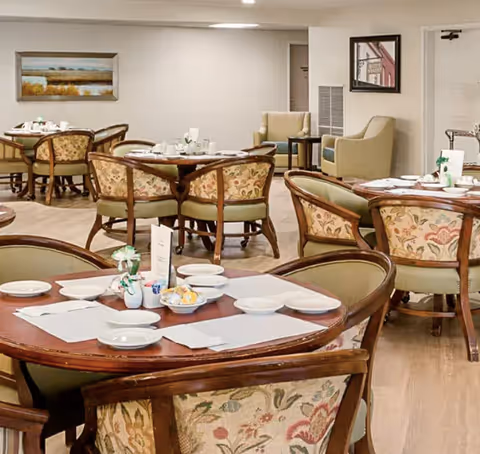 Dining room with round wooden tables set with white plates, bowls, and napkins. Upholstered chairs with floral patterns surround the tables. The room has light-colored walls, a wooden floor, framed artwork on the walls, and a beige armchair in the background.