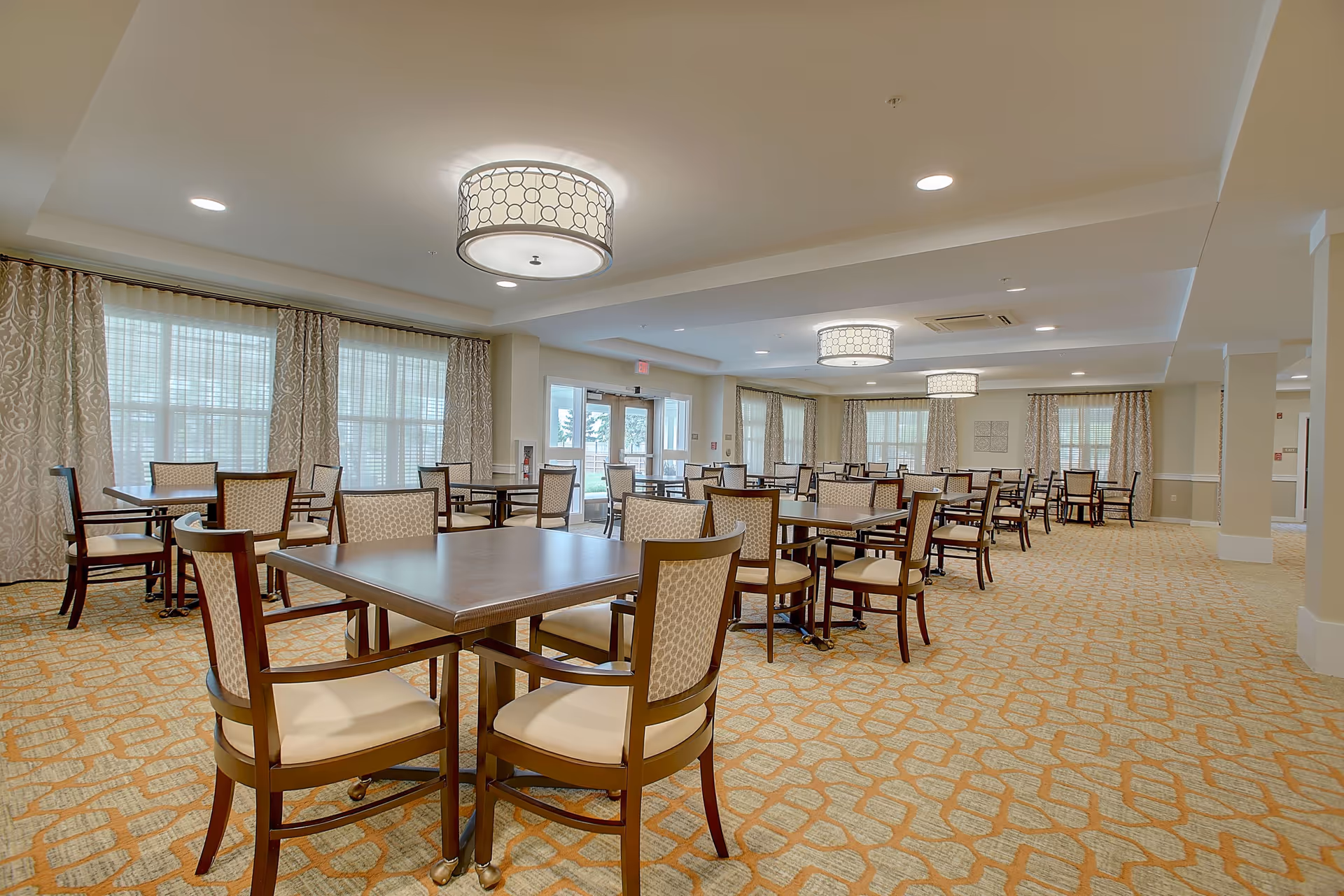 Spacious dining room with multiple wooden tables and cushioned chairs arranged neatly. The room has large windows with patterned curtains allowing natural light to fill the space. The ceiling features modern circular light fixtures, and the floor is carpeted with a geometric pattern.