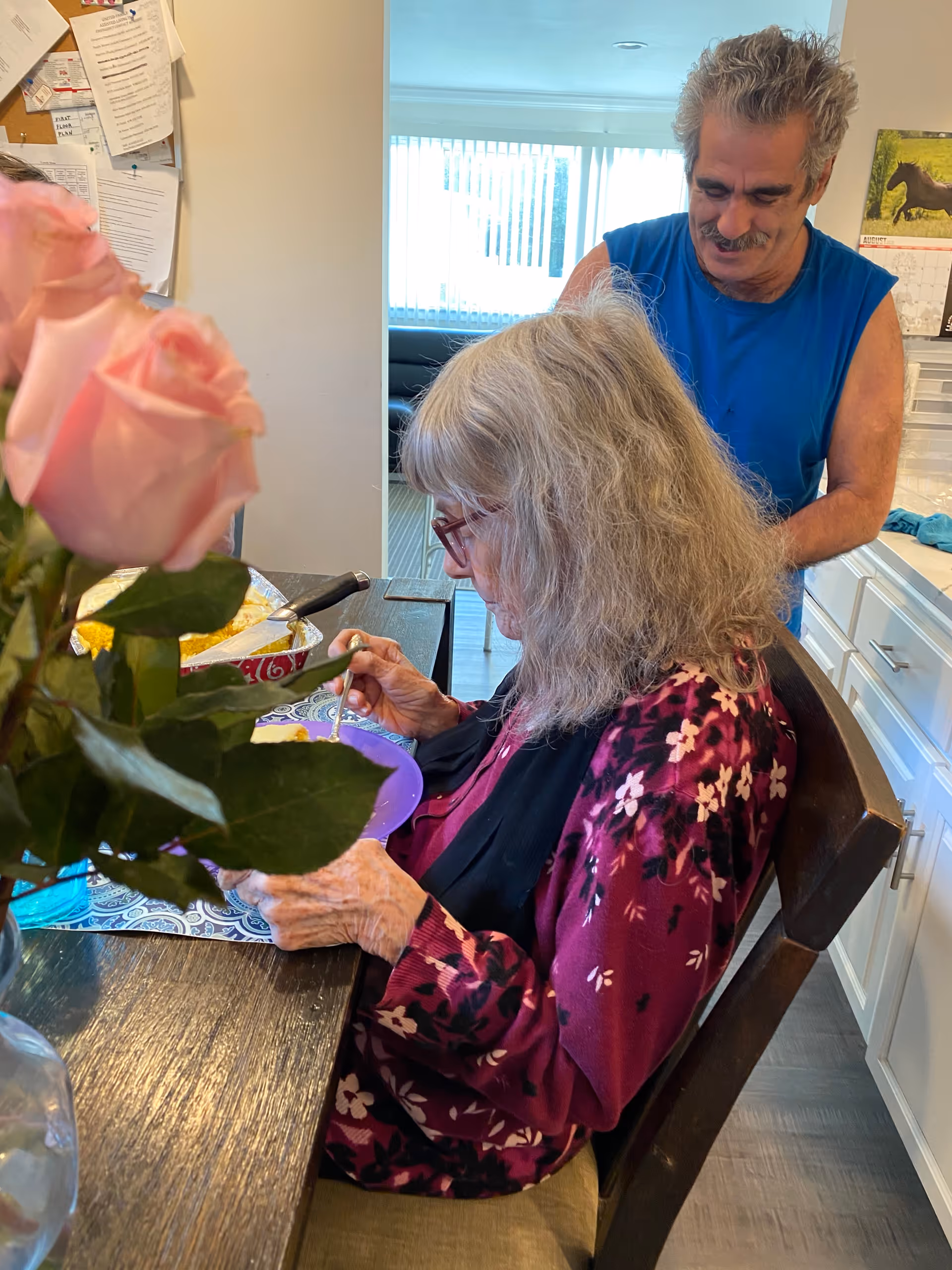 An elderly woman with gray hair and glasses is seated at a dining table eating from a purple plate. A man in a blue sleeveless shirt stands behind her, smiling. There is a vase with pink roses on the table and a kitchen area in the background with white cabinets and a calendar on the wall.