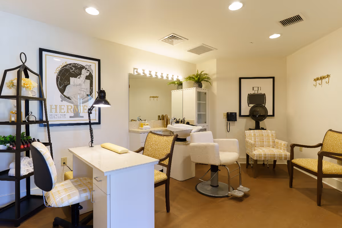 A well-lit salon room with a manicure table and chair on the left, a hair washing station with a white salon chair in the center, and several upholstered chairs arranged around the room. The walls are decorated with framed artwork, and there is a black shelving unit with various items on the left side. The floor is brown, and the ceiling has recessed lighting.