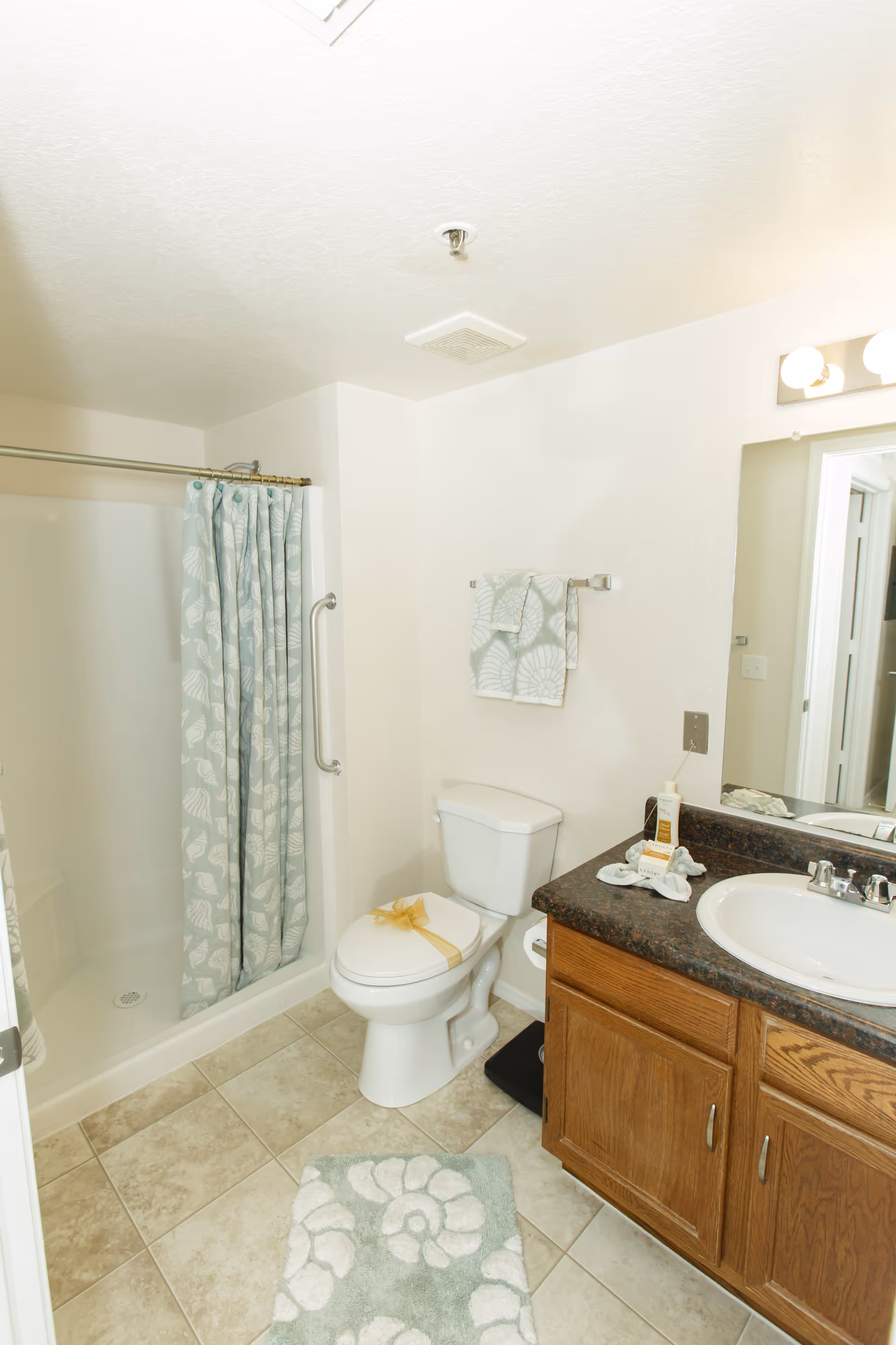 A clean bathroom with a shower and a curtain featuring a seashell pattern, a toilet with a decorative ribbon on the seat, a towel rack with matching seashell patterned towels, a sink with a dark countertop, wooden cabinets below, and a large mirror above the sink.