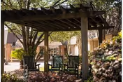 Outdoor seating area at a senior living facility with wooden pergola providing shade over several green chairs. Trees and bushes surround the area, and a building with windows is visible in the background.