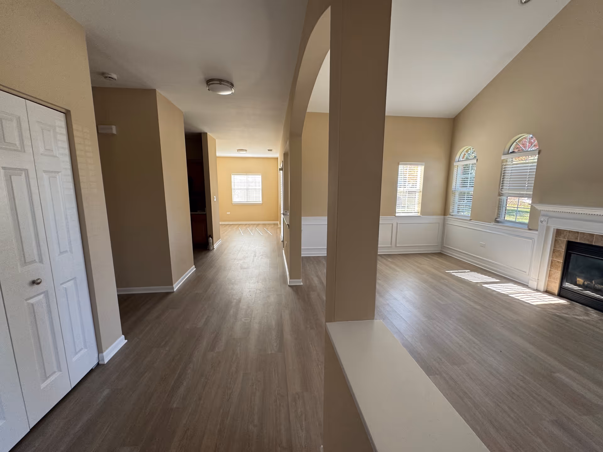 Interior view of a senior living facility showing a hallway leading to a bright room with windows. On the right side, there is a living area with three arched windows, a fireplace, and light wood flooring. The walls are painted beige with white wainscoting.