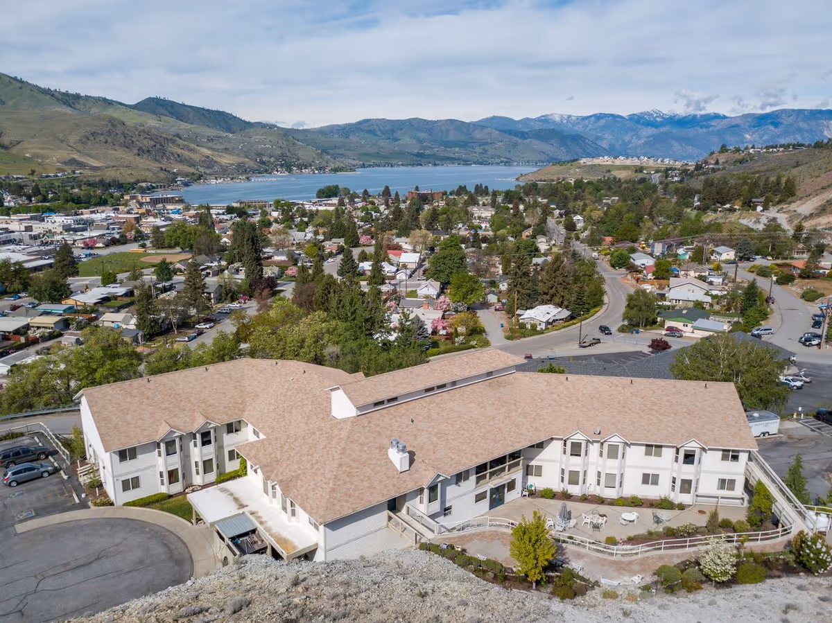 Aerial view of a large senior living facility named Heritage Heights, surrounded by a residential neighborhood with a lake and mountains in the background under a partly cloudy sky.