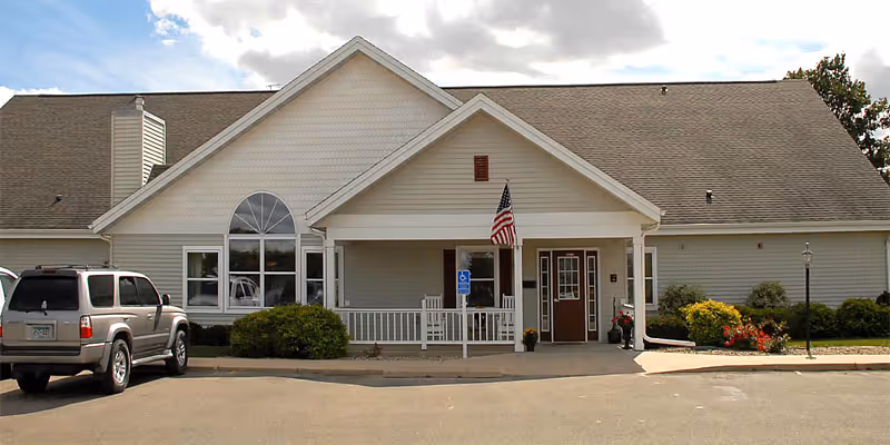 Exterior front view of a single-story senior living facility building with a peaked roof, an American flag near the entrance, a wheelchair accessible sign, and a parked SUV in front.