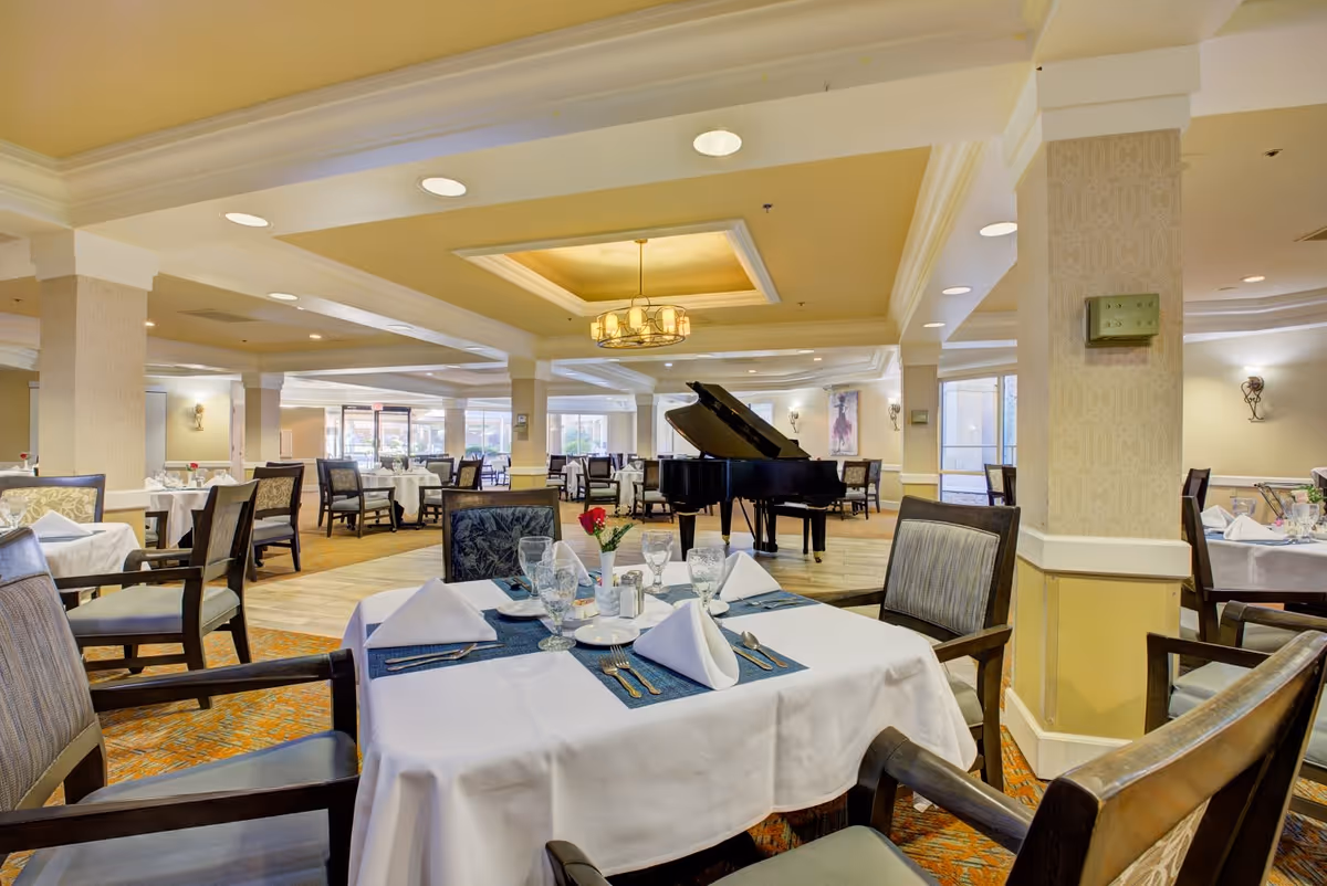 A spacious and well-lit dining room in a senior living facility with multiple tables covered in white tablecloths, set with glassware, napkins, and cutlery. A grand piano is positioned in the center of the room, and large windows allow natural light to fill the space. The room features beige walls, carpeted floors, and elegant ceiling lighting.
