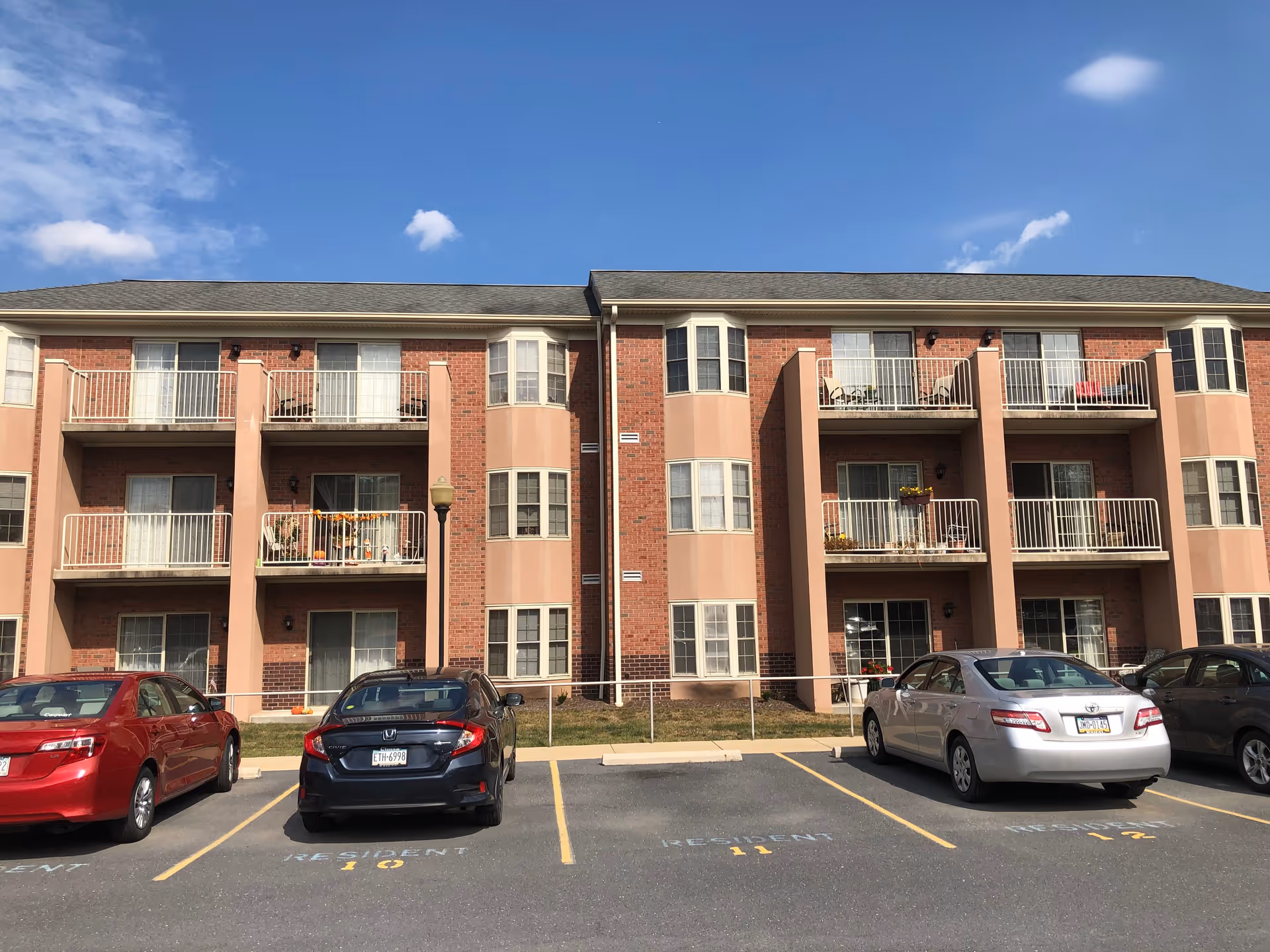 Exterior view of a three-story brick apartment building with balconies and several parked cars in front under a blue sky with a few clouds.