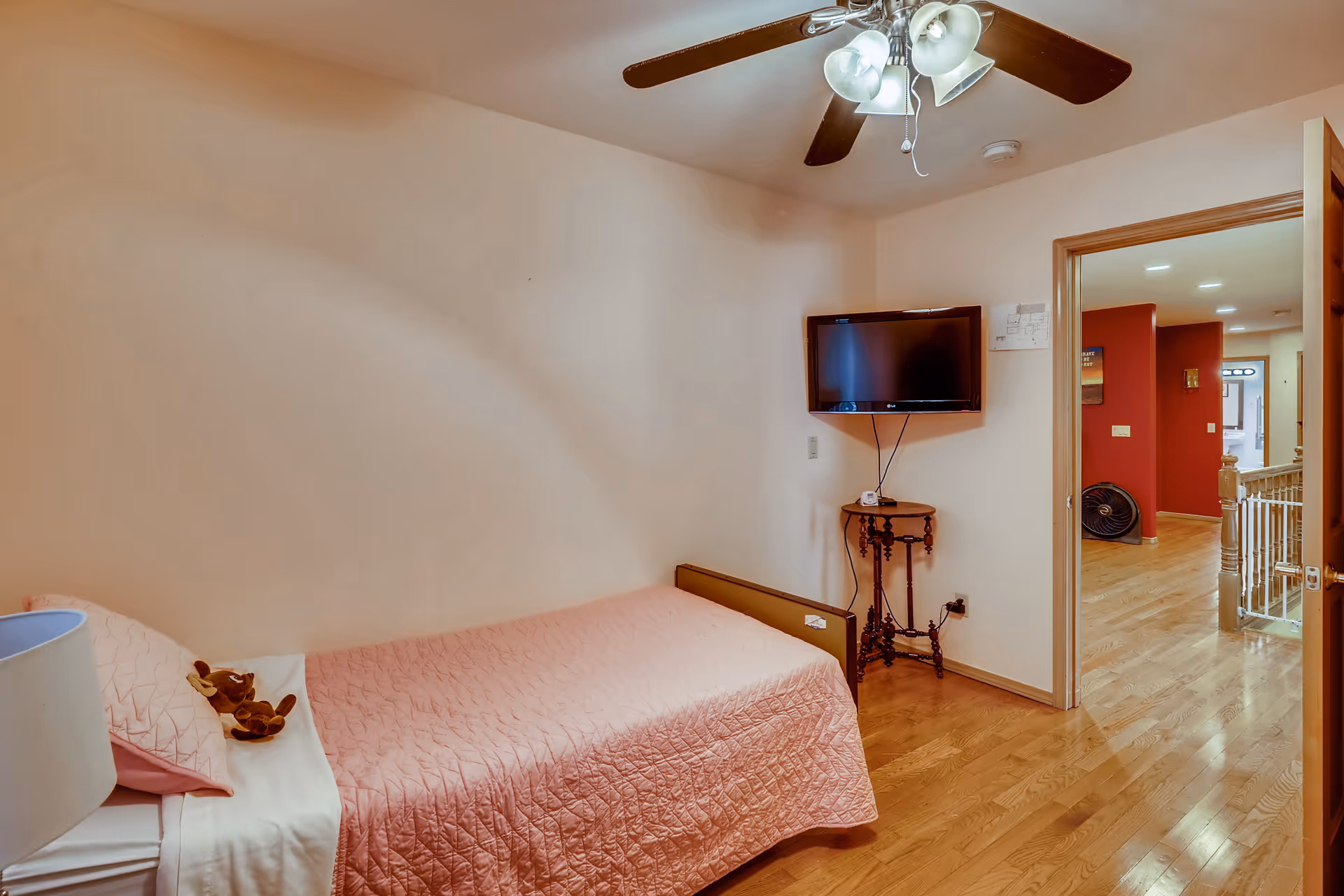 A small bedroom with a single bed covered in a pink quilt and a stuffed animal on the pillow. There is a ceiling fan with lights above, a wall-mounted flat-screen TV, and a small wooden side table beneath the TV. The room has wooden flooring and an open door leading to a hallway with red walls and wooden floors.