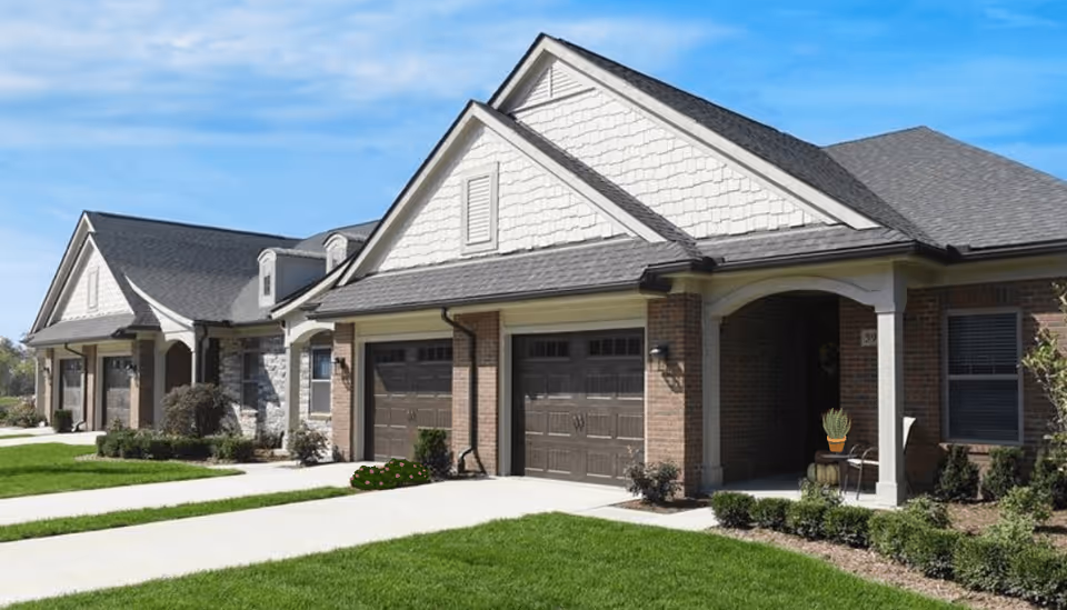 Exterior view of a single-story residential building with brick and siding facade, multiple garages, well-maintained green lawns, shrubs, and a clear blue sky.