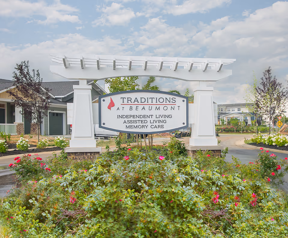 Entrance sign for Traditions at Beaumont senior living facility surrounded by landscaped bushes and flowers, with buildings and a partly cloudy sky in the background.