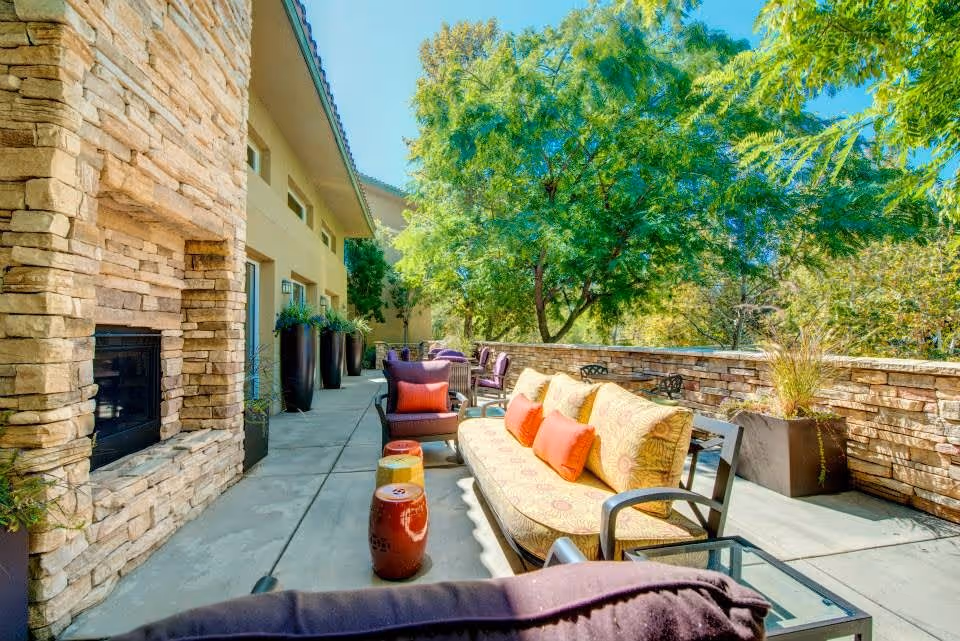 Outdoor patio area with cushioned seating including a sofa and chairs, colorful throw pillows, a stone fireplace on the left, large planters with greenery, and trees in the background under a clear blue sky.