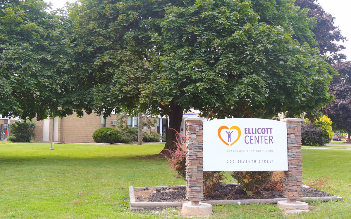 Sign for Ellicott Center on a stone base sits on a grassy lawn under a large tree with the facility building in the background.
