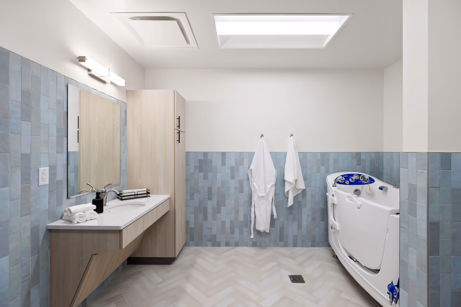 Modern accessible bathroom with a sink and mirror on the left, two towels hanging on the back wall, and a walk-in bathtub on the right against blue-tiled walls.