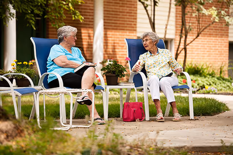 Two elderly women sitting on blue chairs outside in a garden area, engaged in conversation. One woman is wearing a light blue shirt and black shorts, holding a book, while the other is wearing a floral blouse and white pants with a red handbag placed on the ground beside her. There are potted plants on a small table between them and a brick building in the background.