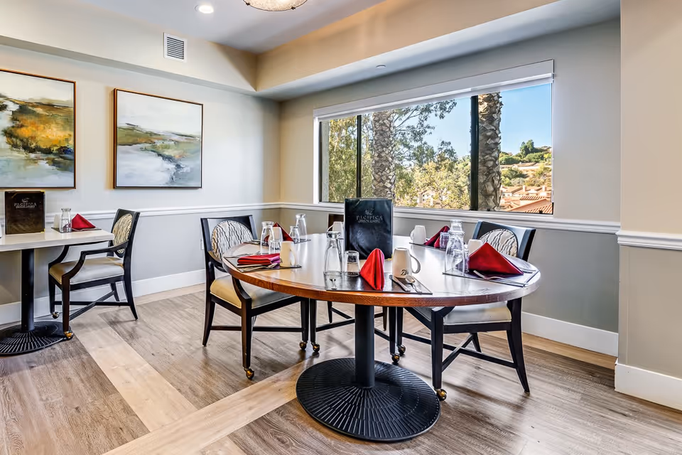 A bright dining room with a round wooden table set for four people, featuring red folded napkins, glasses, and menus. The room has large windows showing trees and rooftops outside, light-colored walls with two abstract paintings, and wooden flooring.