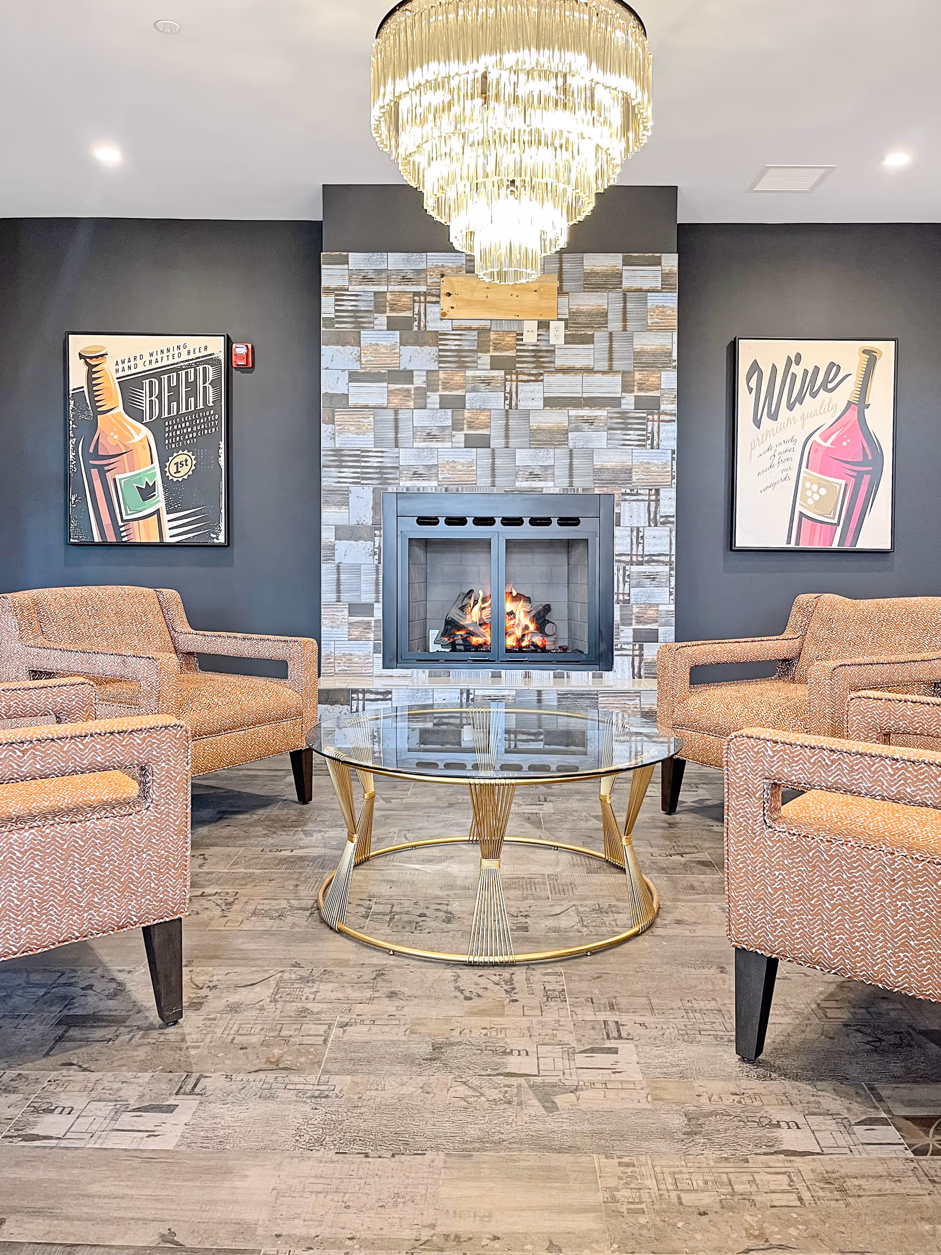 A cozy lounge area with four patterned armchairs arranged around a round glass coffee table with a gold metal frame. Behind the seating is a modern fireplace with a tiled surround and a fire burning inside. Above the fireplace hangs a large, elegant chandelier. On the dark gray walls on either side of the fireplace are framed posters, one featuring a beer bottle and the other a wine bottle.