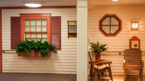 Indoor area designed to resemble an exterior porch with beige siding walls, a window with red shutters and a flower box filled with green plants, a decorative octagonal window, a wall-mounted light fixture, and two wooden chairs with cushions next to a small table holding a potted plant.