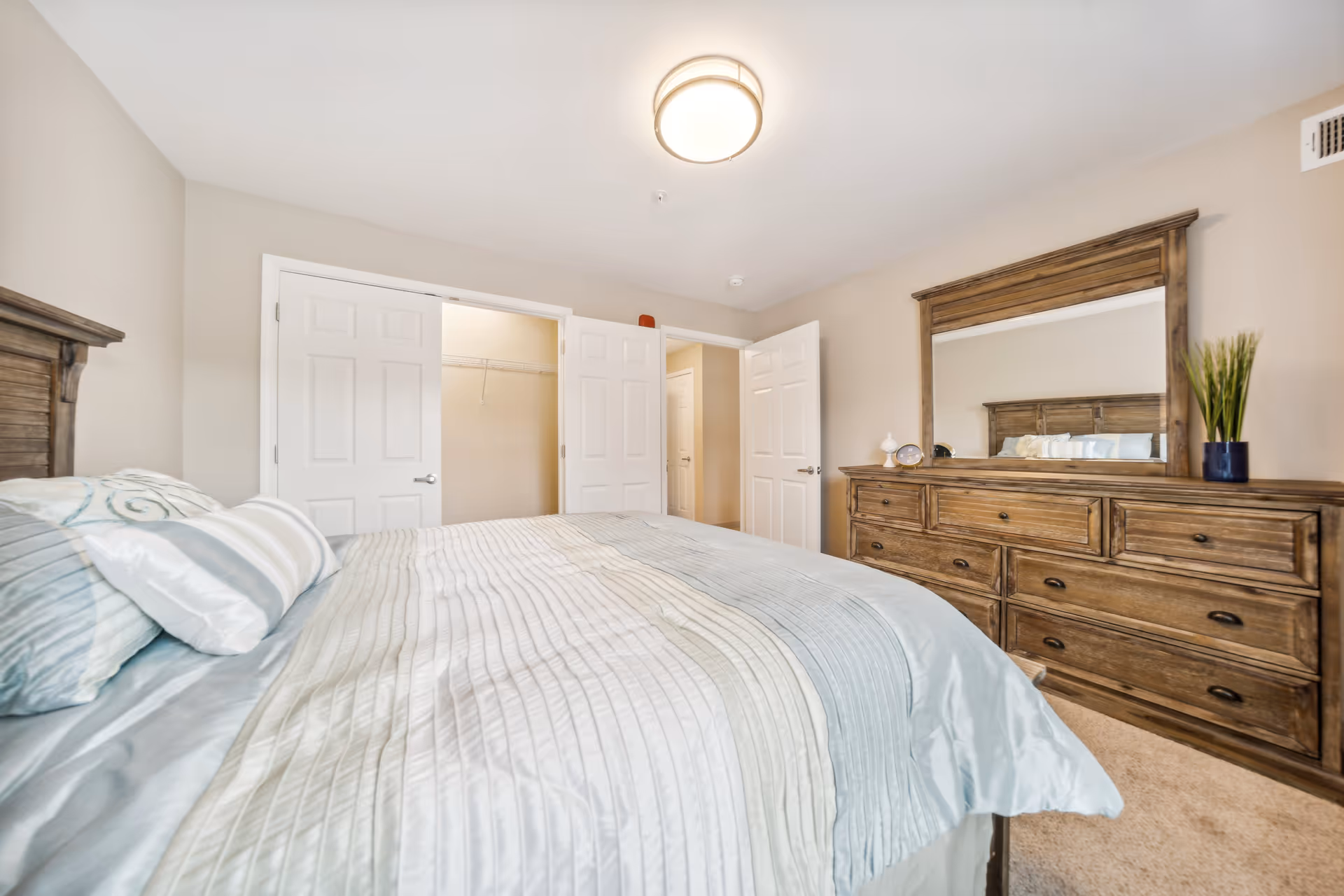 A bedroom with a large bed covered in a striped comforter in shades of blue, beige, and white. The room features a wooden dresser with a large mirror, decorated with a small clock, a white decorative item, and a potted plant. There are two open white doors, one leading to a closet and the other to a hallway. The walls are painted light beige and the ceiling has a round light fixture.