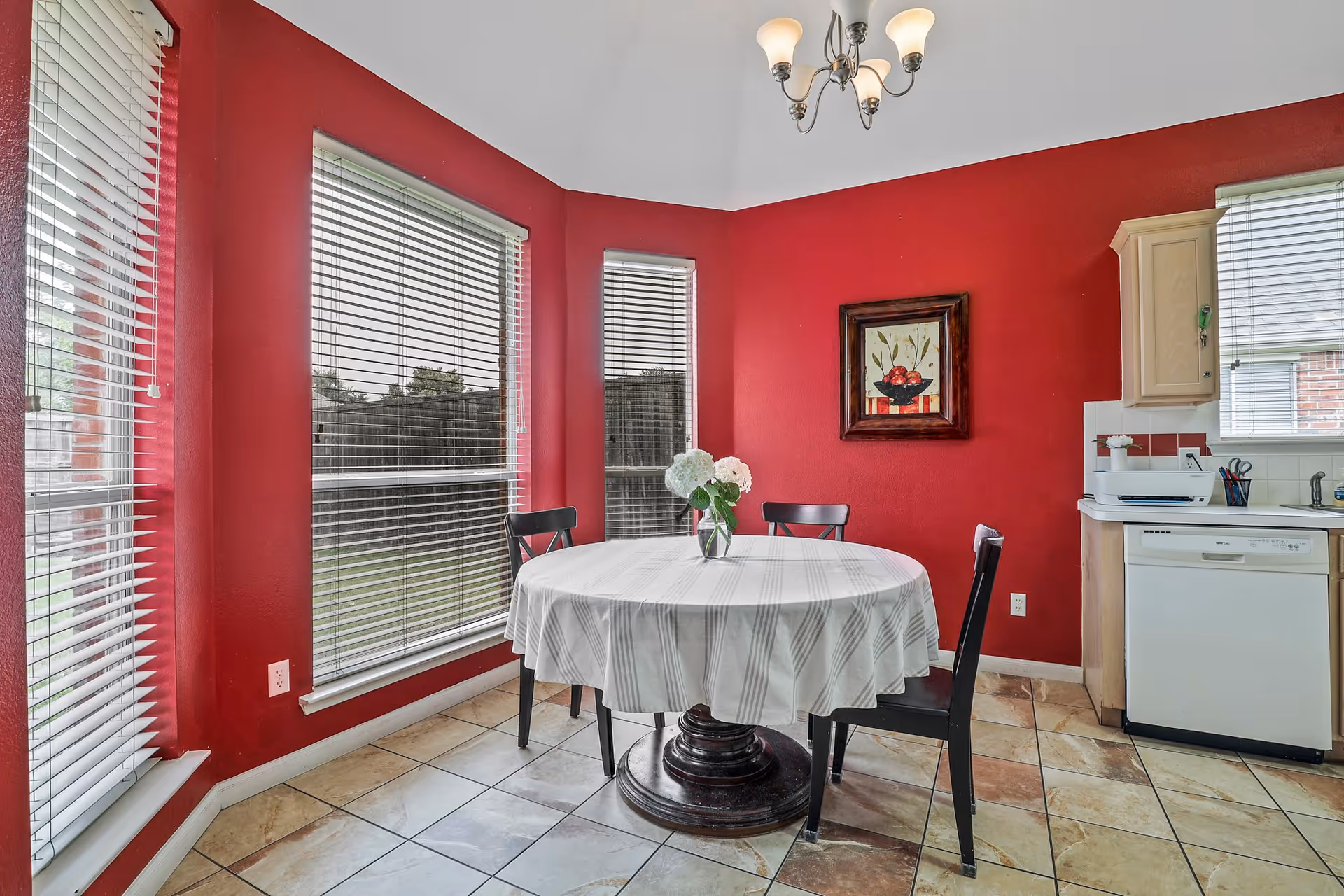 A dining area with red walls featuring three large windows with blinds. There is a round table covered with a white tablecloth and a vase with white flowers in the center. Four black chairs surround the table. A framed painting hangs on the wall, and part of a kitchen with a dishwasher and countertop is visible on the right side. The floor is tiled, and a chandelier with three lights hangs from the ceiling.