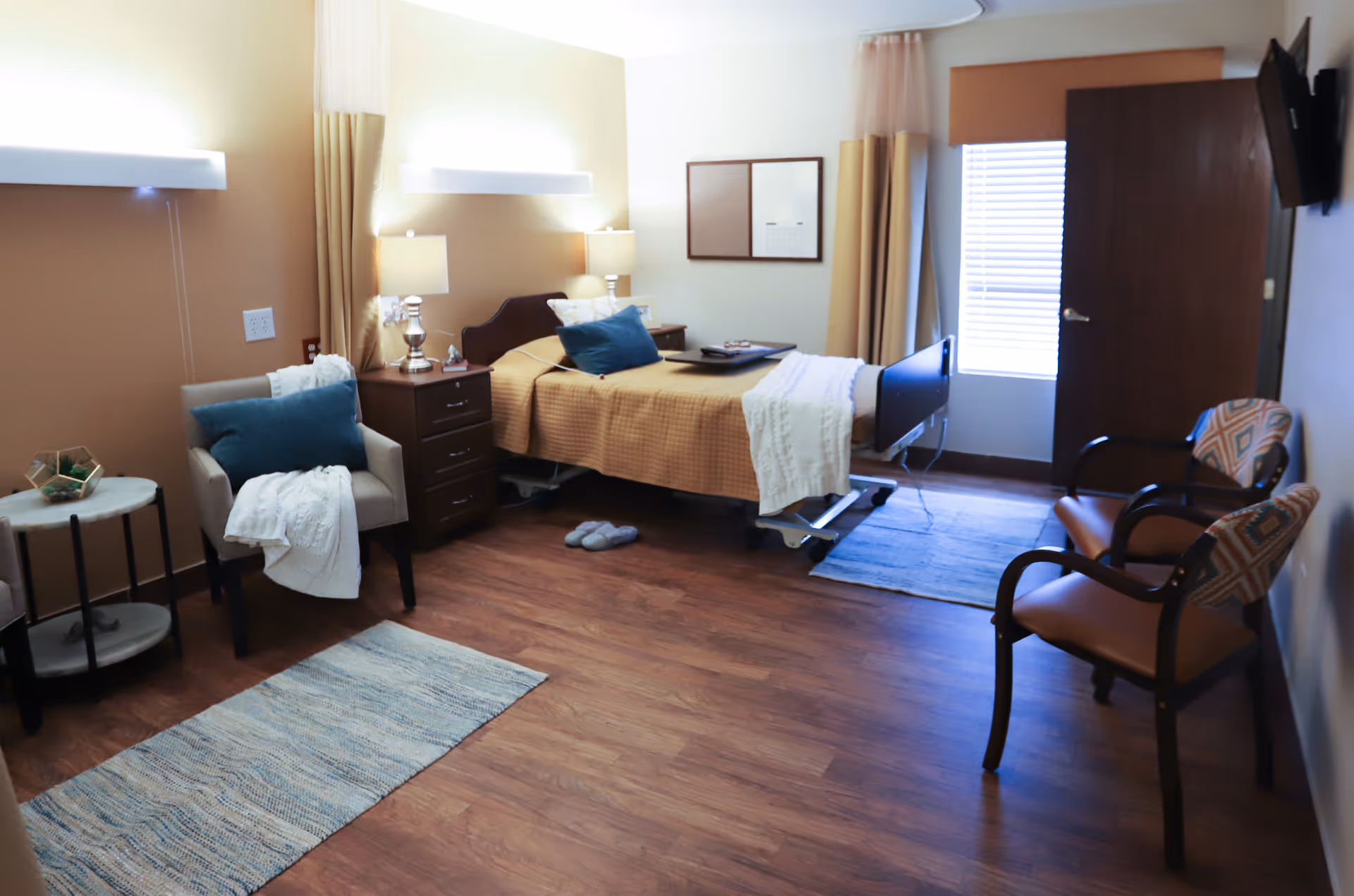 A well-lit nursing facility room with a single bed covered in a beige blanket and white throw, a bedside table with a lamp, a comfortable armchair with a blue cushion and white throw, two wooden chairs with patterned cushions, a wall-mounted TV, and a window with blinds and beige curtains. The floor is wooden with two blue rugs.